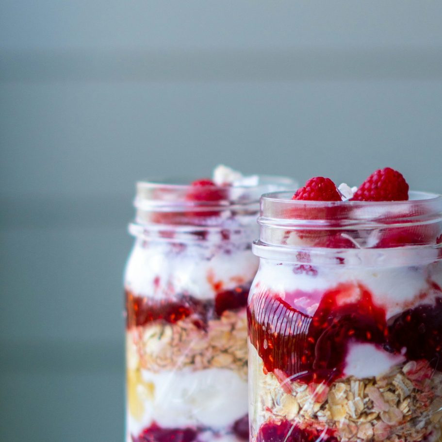 Zwei Gläser mit Schichten aus Joghurt, Beeren und Müsli, dekoriert mit Himbeeren.